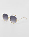 A pair of Drape sunglasses by For Art's Sake® with gold frames and dark, gradient lenses. The design features a unique, slightly wavy cut on the top edges of the oversized squared frame. Intricate metal detailing adds to their charm as they rest on a white surface, viewed at an angle.