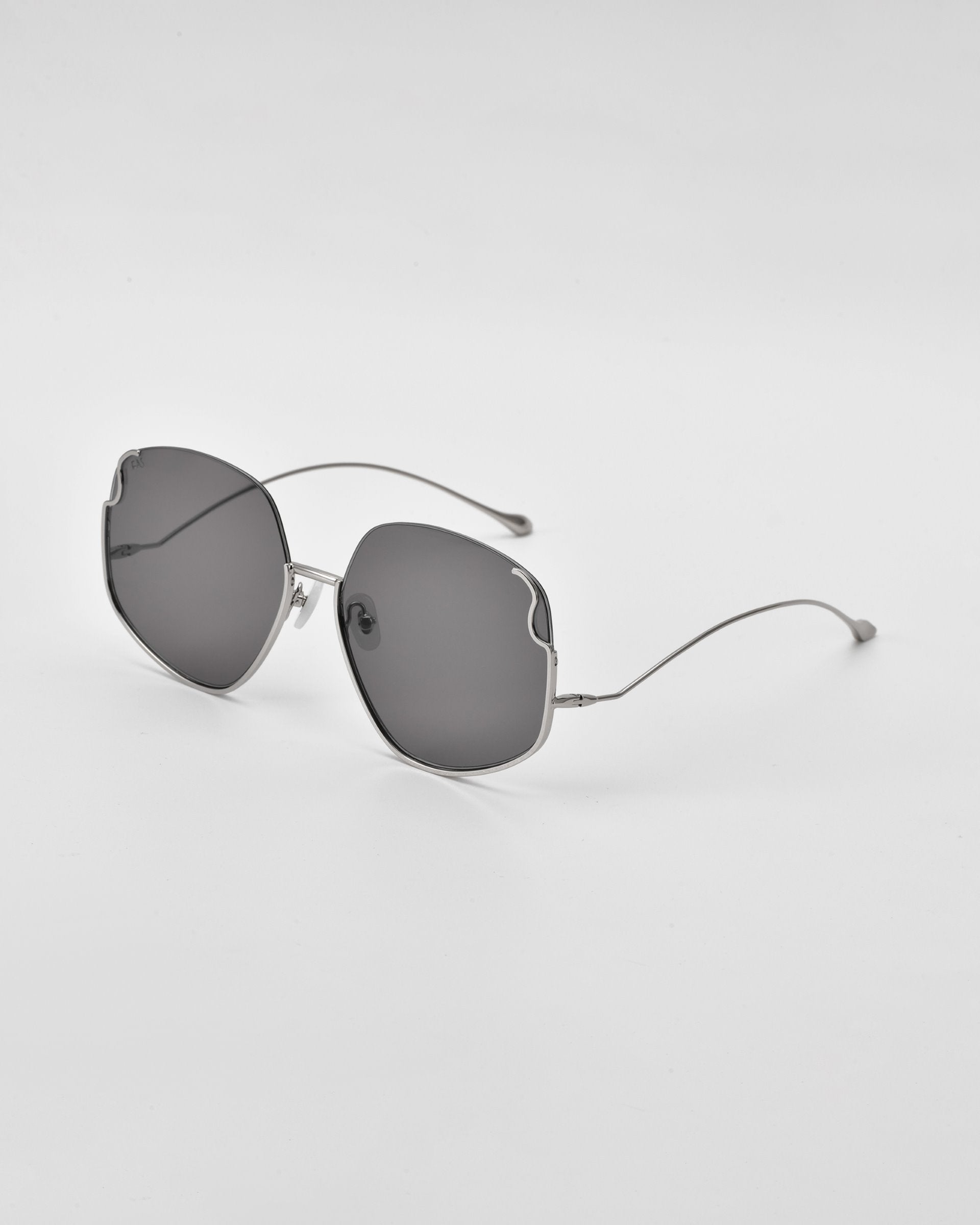 A pair of Drape sunglasses by For Art&#39;s Sake® with curved, wireframe arms and large, slightly rounded, dark lenses. The frames are fine and metallic with intricate metal detailing, creating a modern and minimalist look. The glasses are placed on a plain, light-colored surface.