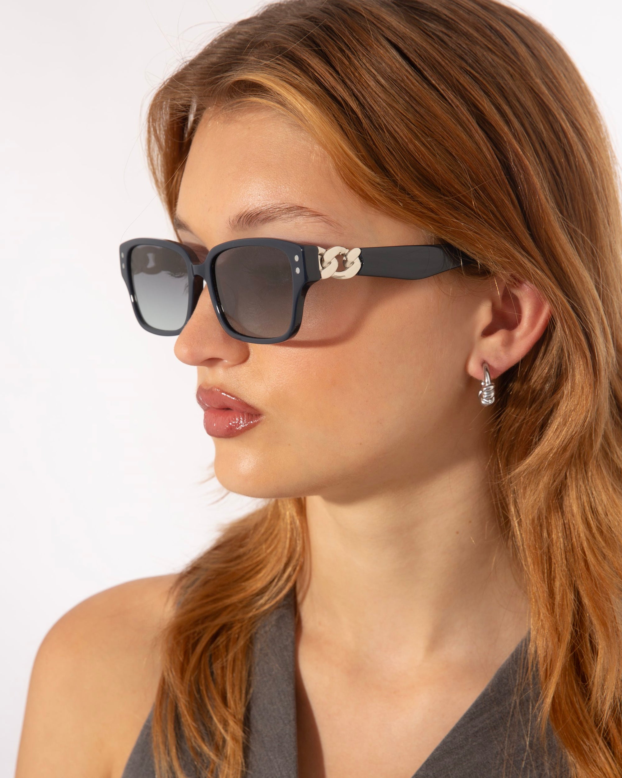 A person with long, light brown hair is wearing black Zenith sunglasses by For Art&#39;s Sake® with a small decorative chain on the sides, capturing &#39;90s styles. They have a pair of drop earrings and are looking slightly to the left. The background is plain white.
