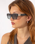 A person with long, light brown hair is wearing black Zenith sunglasses by For Art's Sake® with a small decorative chain on the sides, capturing '90s styles. They have a pair of drop earrings and are looking slightly to the left. The background is plain white.