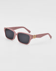 The For Art's Sake® Zenith sunglasses are a pair of pink rectangular sunglasses with dark lenses. Handcrafted from acetate, the frames feature a small, chunky chain detailing in gold on both sides near the temples, adding a decorative element against the white background.