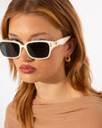 A person with light brown hair wears stylish, white-framed Zenith handcrafted acetate sunglasses with black lenses and gold accents by For Art's Sake®, reminiscent of '90s styles. They are also wearing gold hoop earrings and a beige top. The background is plain white.