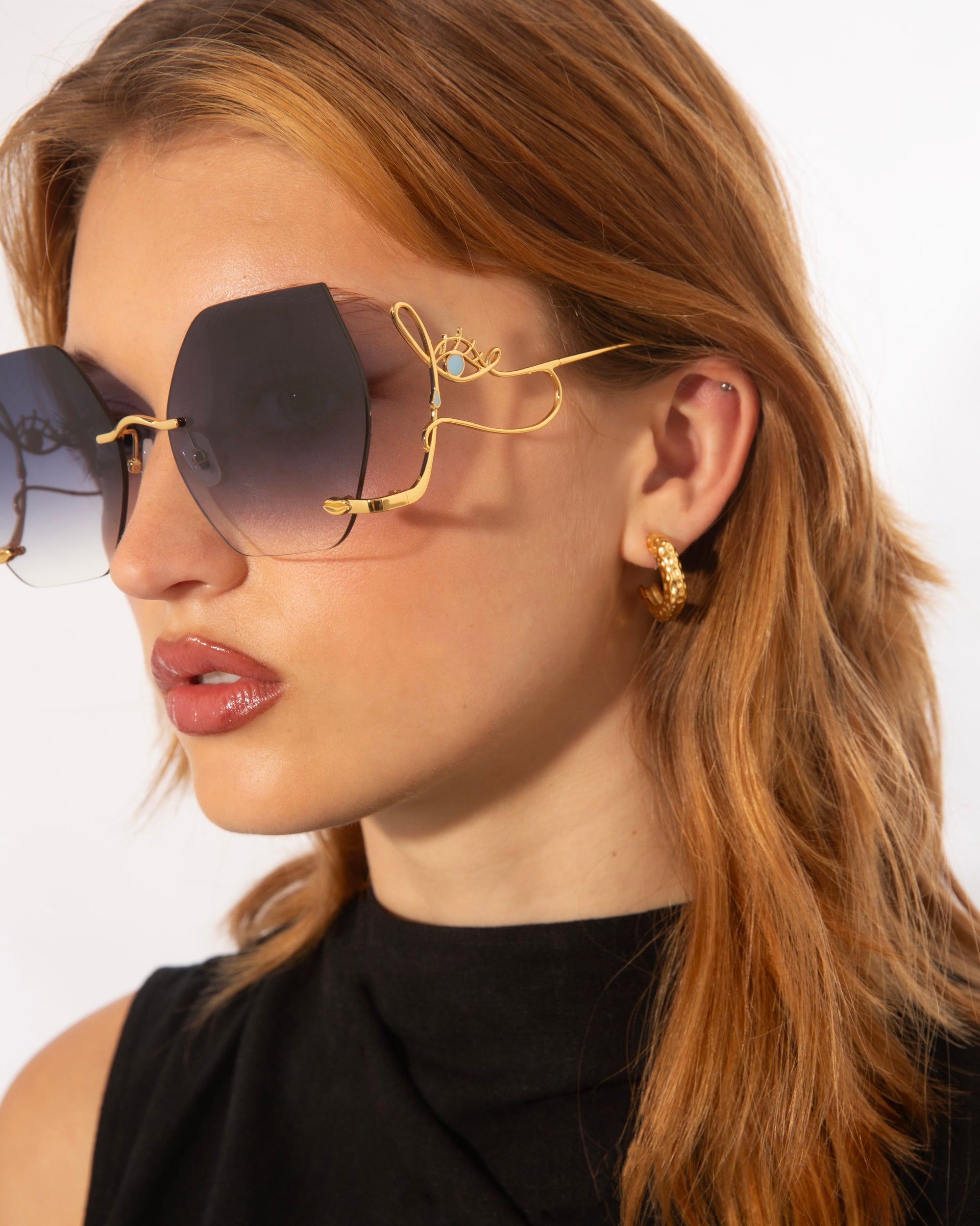 A woman with light brown hair wears large, hexagonal Cry Me a River sunglasses from For Art's Sake® with gradient lenses and gold wireframes. She has a small hoop earring and is dressed in a sleeveless black top, looking to the left against a plain white background. This limited edition accessory exudes timeless elegance.
