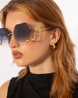 A woman with light brown hair wears large, hexagonal Cry Me a River sunglasses from For Art's Sake® with gradient lenses and gold wireframes. She has a small hoop earring and is dressed in a sleeveless black top, looking to the left against a plain white background. This limited edition accessory exudes timeless elegance.