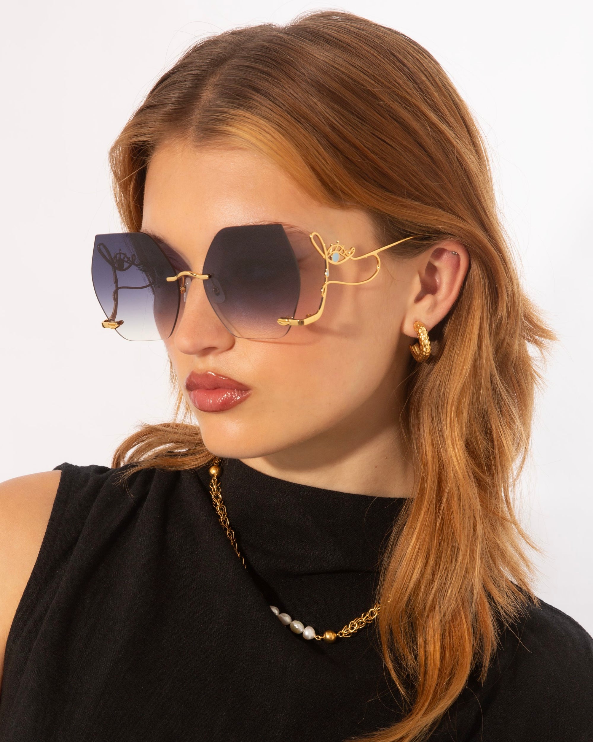 A woman with light brown hair wears oversized, gradient Cry Me a River sunglasses with gold detailing. She is dressed in a black top and accessorizes with hoop earrings and a gold chain necklace. Like Julie London&#39;s song, this limited edition style by For Art&#39;s Sake® exudes timeless elegance as she looks to her left against a plain white background.