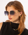 A woman with light brown hair wears oversized, gradient Cry Me a River sunglasses with gold detailing. She is dressed in a black top and accessorizes with hoop earrings and a gold chain necklace. Like Julie London's song, this limited edition style by For Art's Sake® exudes timeless elegance as she looks to her left against a plain white background.