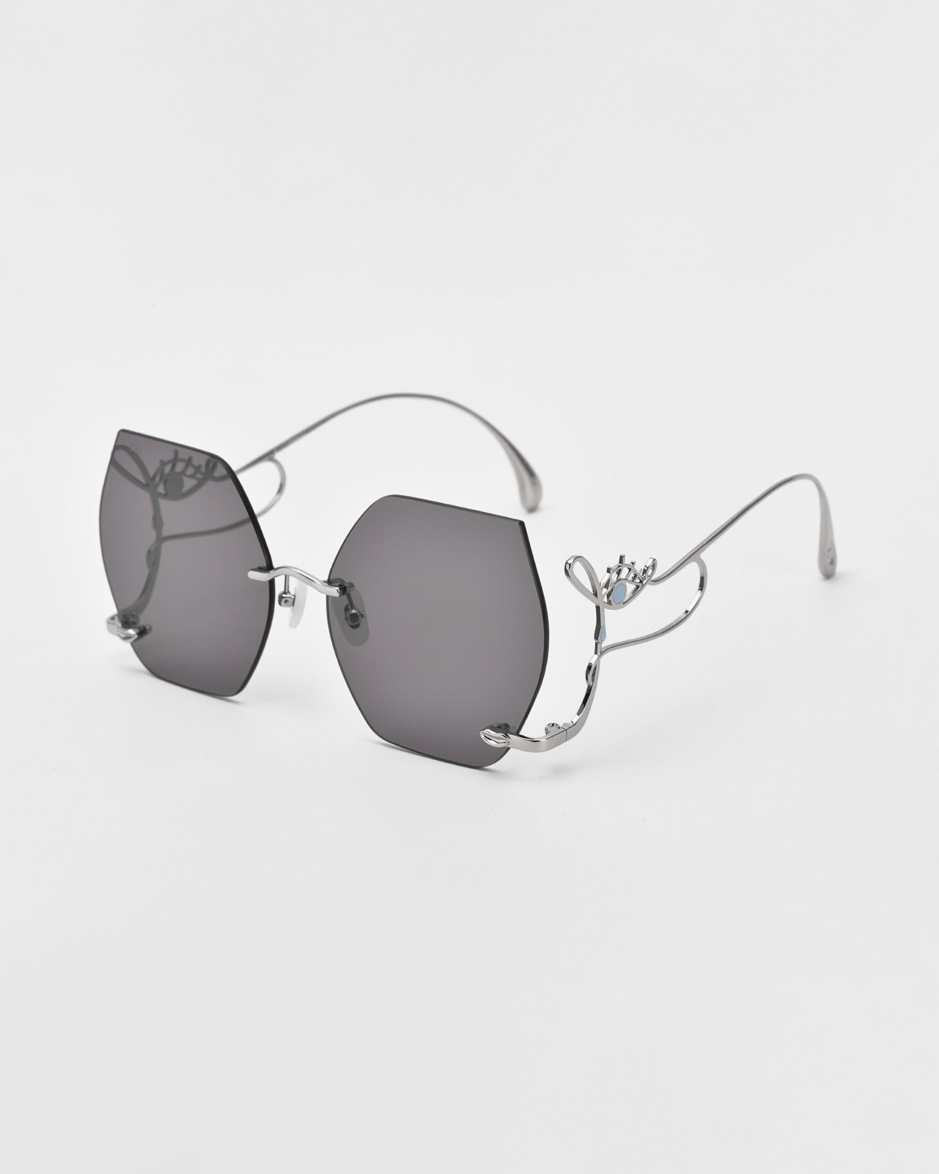 A pair of stylish, limited edition Cry Me a River sunglasses by For Art&#39;s Sake® with hexagonal-shaped dark-tinted lenses and thin silver metal frames. The design features an intricate decorative element on the arms, including a small crown detail and a tiny blue gem. The background is solid white.
