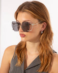 A person with long, light brown hair is wearing stylish, oversized square sunglasses with intricate wire detailing on the frames. These limited edition statement sunglasses, known as For Art's Sake® "Cry Me a River" sunglasses, complement their sleeveless grey top and small dangling earrings against a plain white wall backdrop.