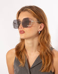 A person with long, wavy, light brown hair is shown from the shoulders up, gazing to the side. They are wearing large Cry Me a River sunglasses from For Art's Sake® with dark lenses and thin silver frames. They are also wearing a sleeveless gray top and drop-style silver earrings.