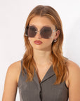 A woman with long, light brown hair is wearing large, limited edition For Art's Sake® Cry Me a River sunglasses with a metallic frame. She has a serious expression and is dressed in a gray sleeveless top. The background is plain white.