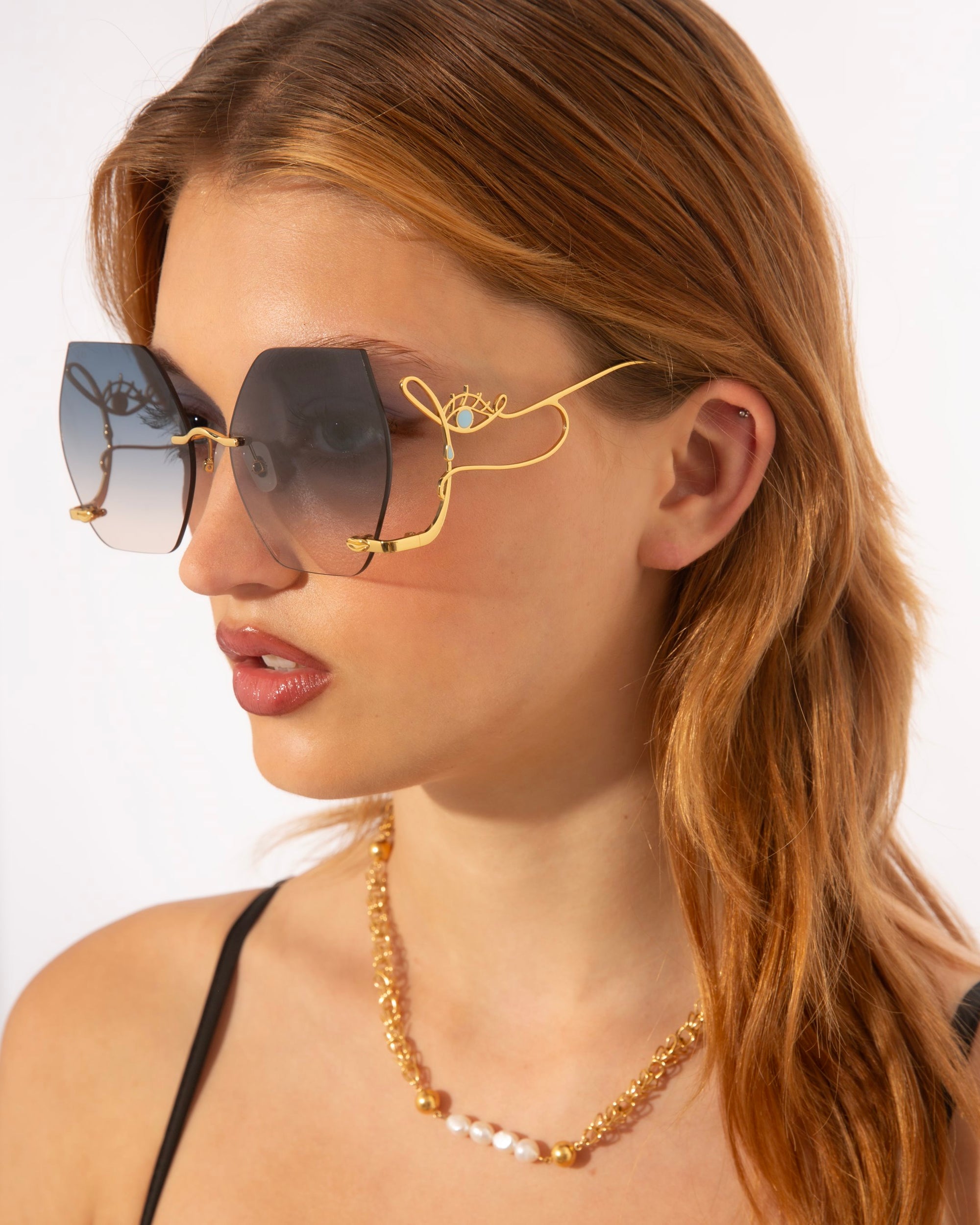 A woman with light brown hair is wearing large, geometric-shaped For Art&#39;s Sake® Cry Me a River statement sunglasses with gradient lenses and decorative gold wire detailing on the frames. She is also wearing a gold chain necklace with pearl accents. The background is plain white.