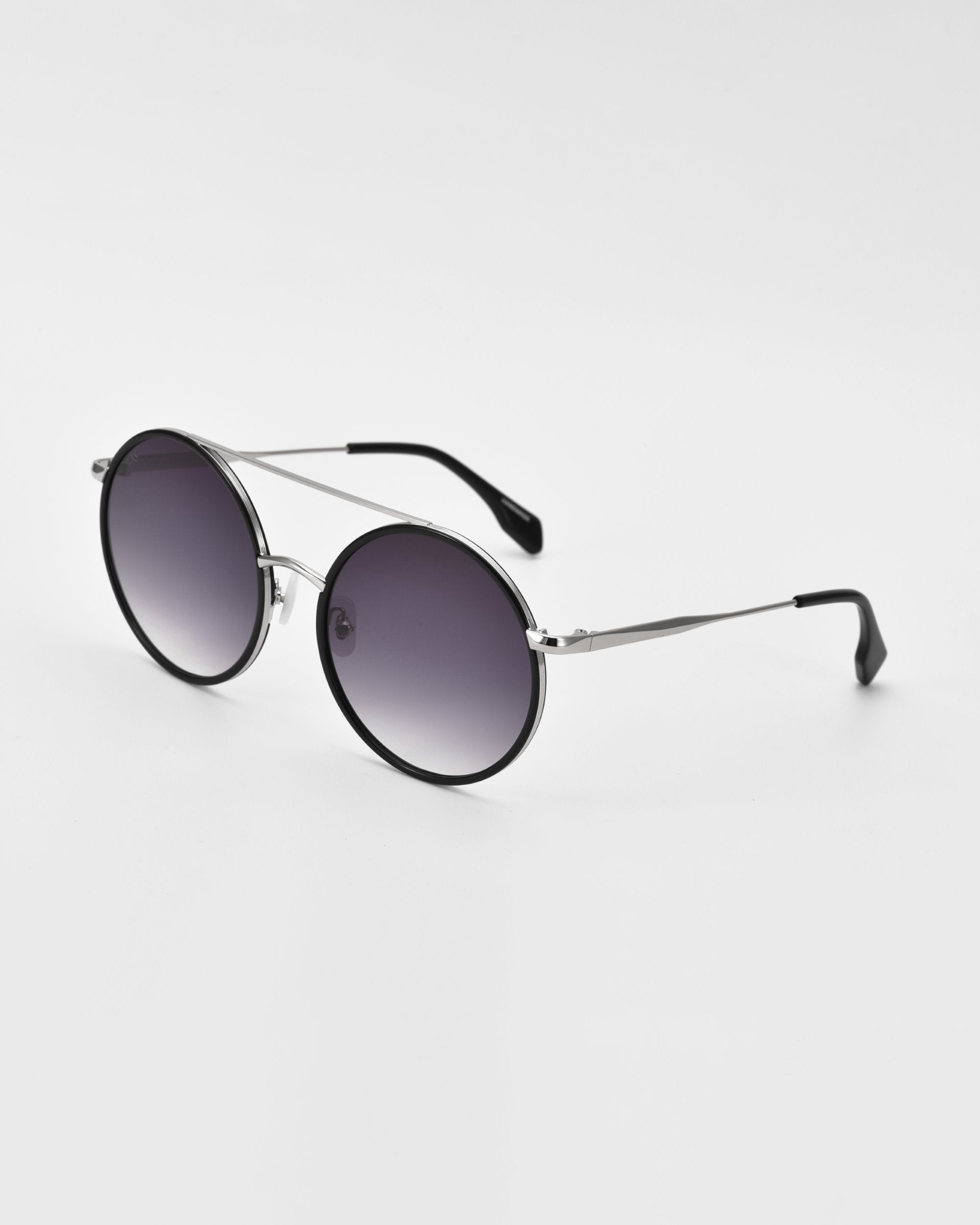 A pair of oversized round aviator sunglasses with black-rimmed, dark gradient lenses and thin, silver arms that have black ear tips. Crafted from gold and silver-tone metal, these stylish shades also feature unique jade-stone nose pads. The For Art&#39;s Sake® Orb sunglasses are placed on a flat, white surface, angled slightly to the right.