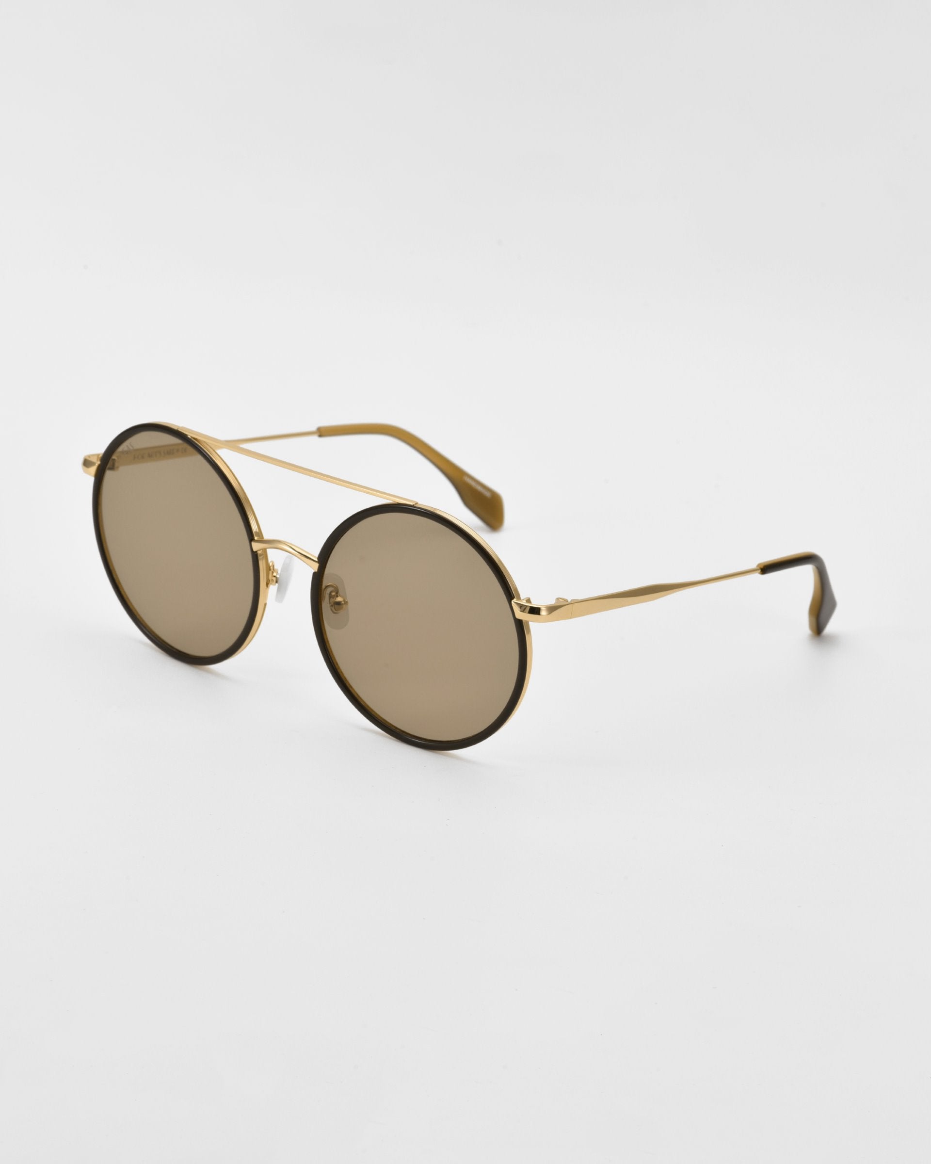 A pair of oversized round aviator sunglasses with gold and silver-tone metal frames and brown lenses. The Orb by For Art&#39;s Sake®, featuring jade-stone nose pads, are placed on a plain white surface, facing slightly to the left with their thin arms extended.