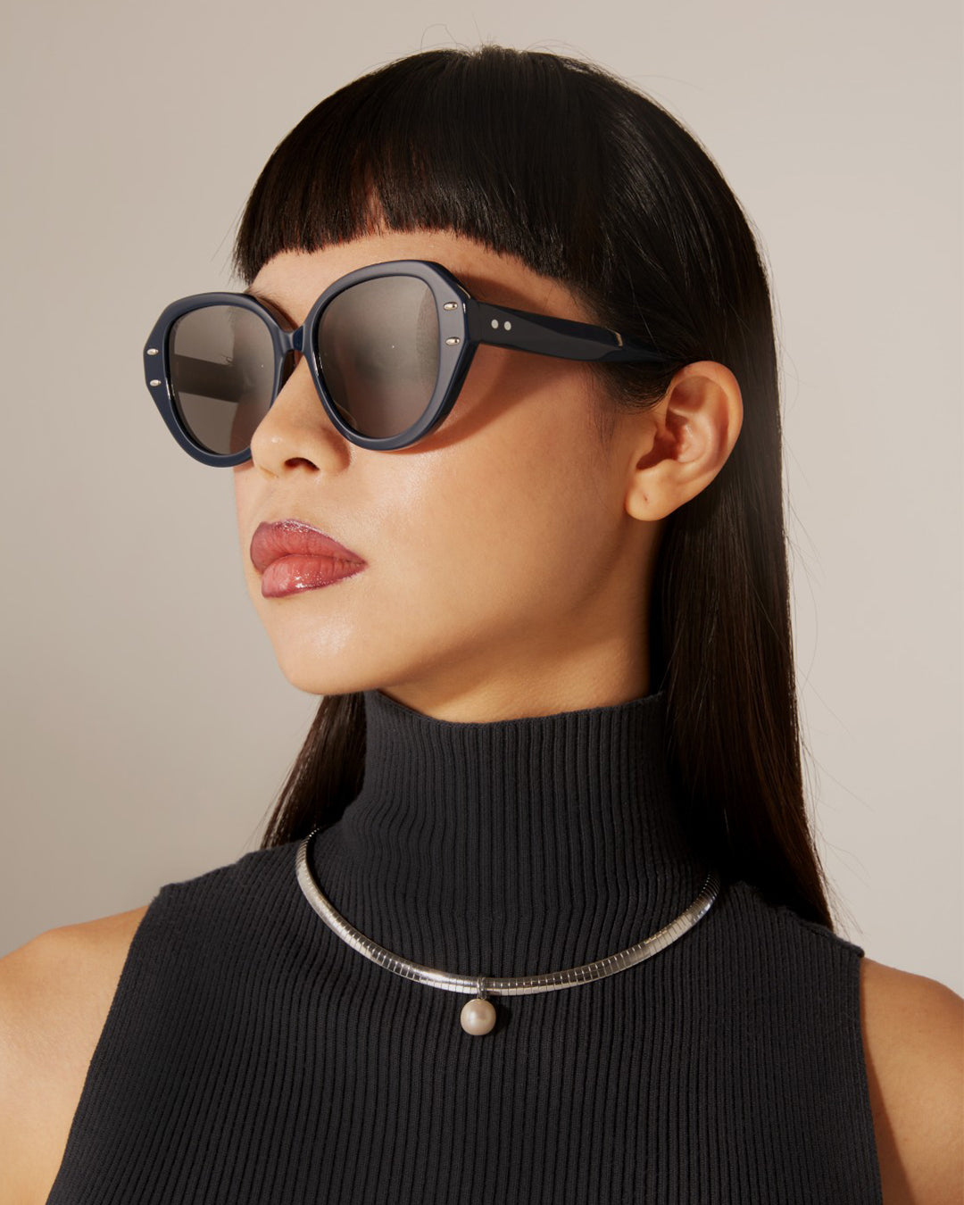 A person with long dark hair and straight bangs wears stylish Mirage sunglasses by For Art's Sake®, a black sleeveless turtleneck top, and a gold and silver-tone metal necklace with a single pearl pendant. The background is plain and light-colored.