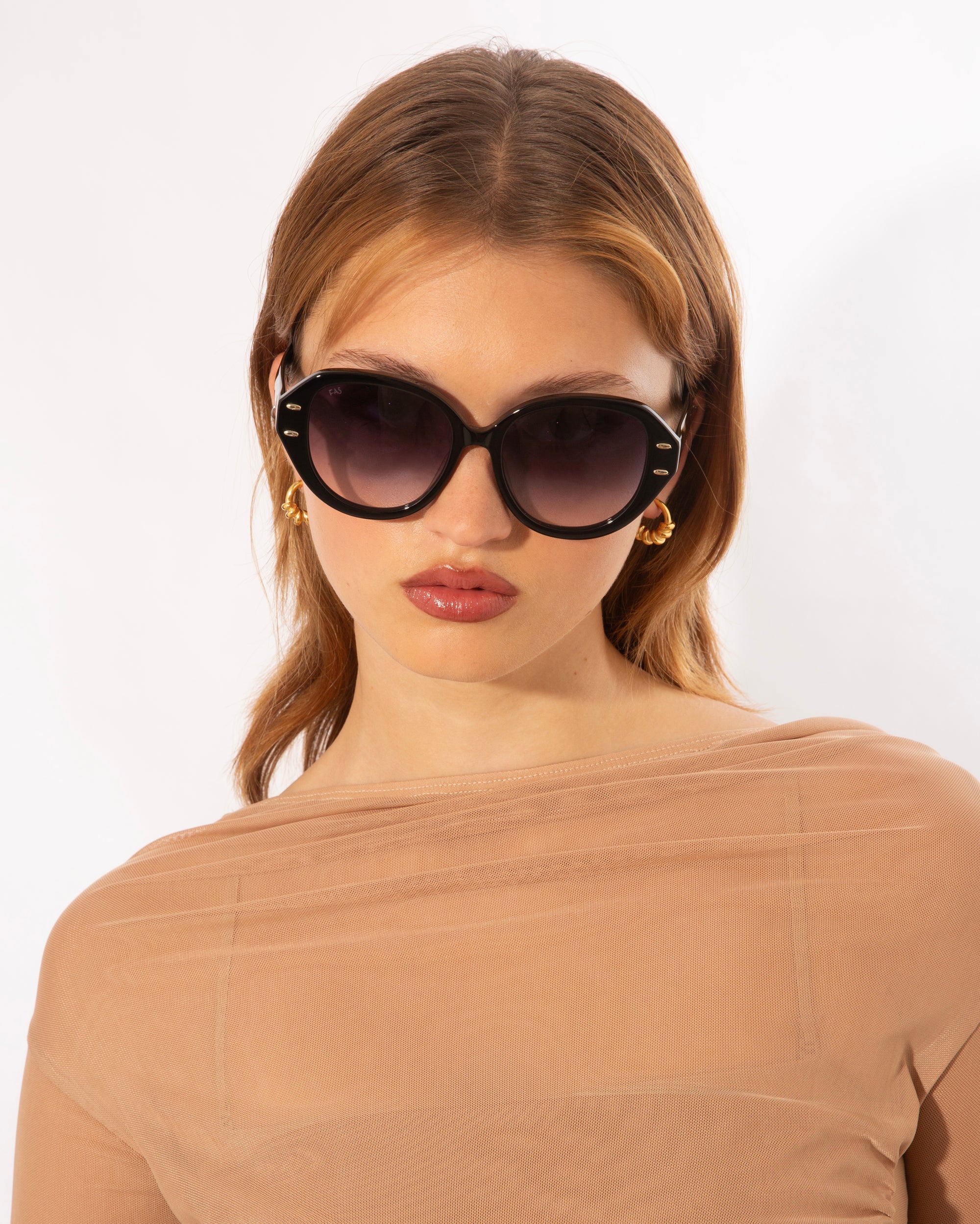 A person with light brown hair wearing large, round Mirage sunglasses by For Art&#39;s Sake® with intricate stud details and gold hoop earrings. They have a neutral expression and are dressed in a sheer, beige top. The background is plain and white.