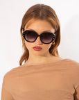 A person with light brown hair wearing large, round Mirage sunglasses by For Art's Sake® with intricate stud details and gold hoop earrings. They have a neutral expression and are dressed in a sheer, beige top. The background is plain and white.