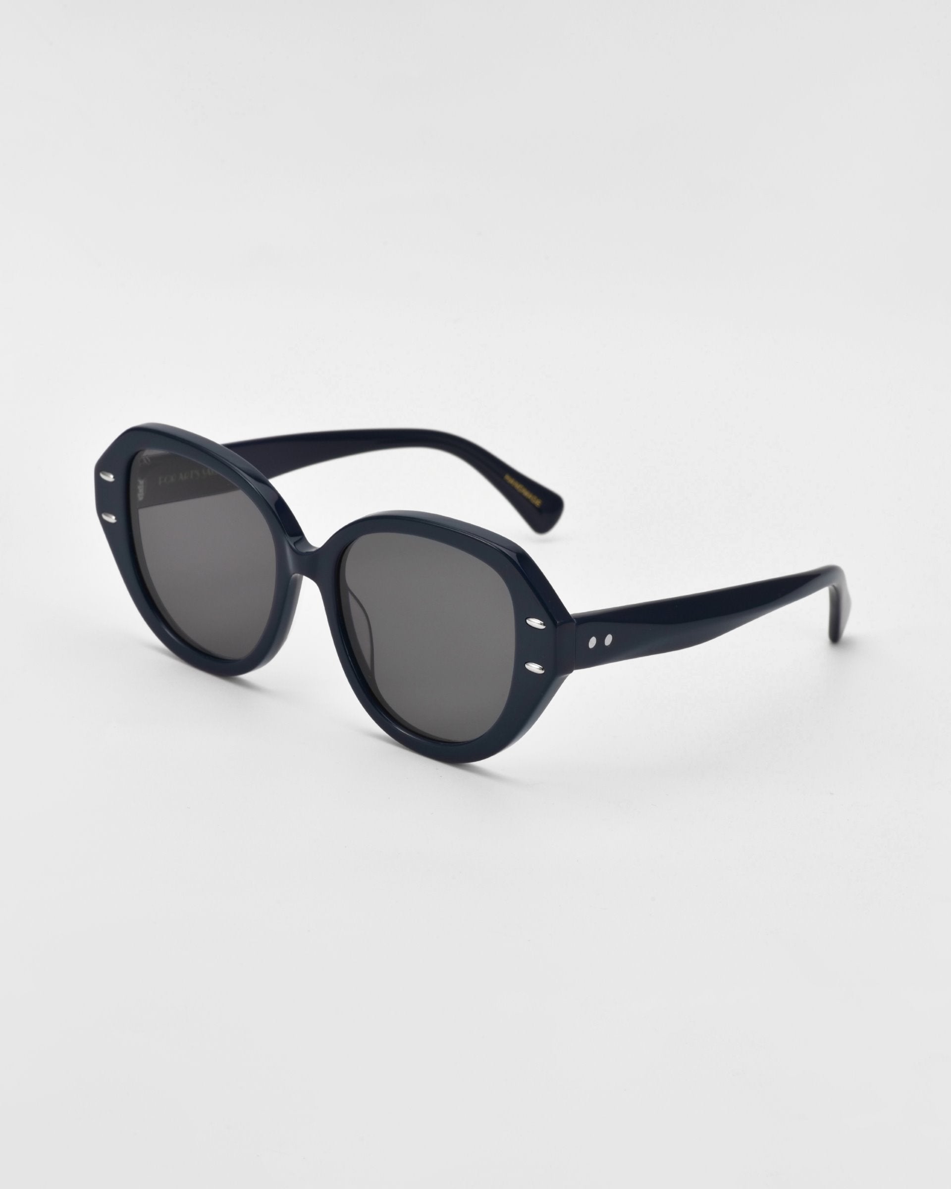 A pair of black, thick-framed cat-eye sunglasses with round lenses and small decorative silver dots near the hinges is positioned on a plain white background. The frames are crafted from plant-based acetate, adding an eco-friendly touch to this stylish accessory. These are the Mirage by For Art's Sake®.