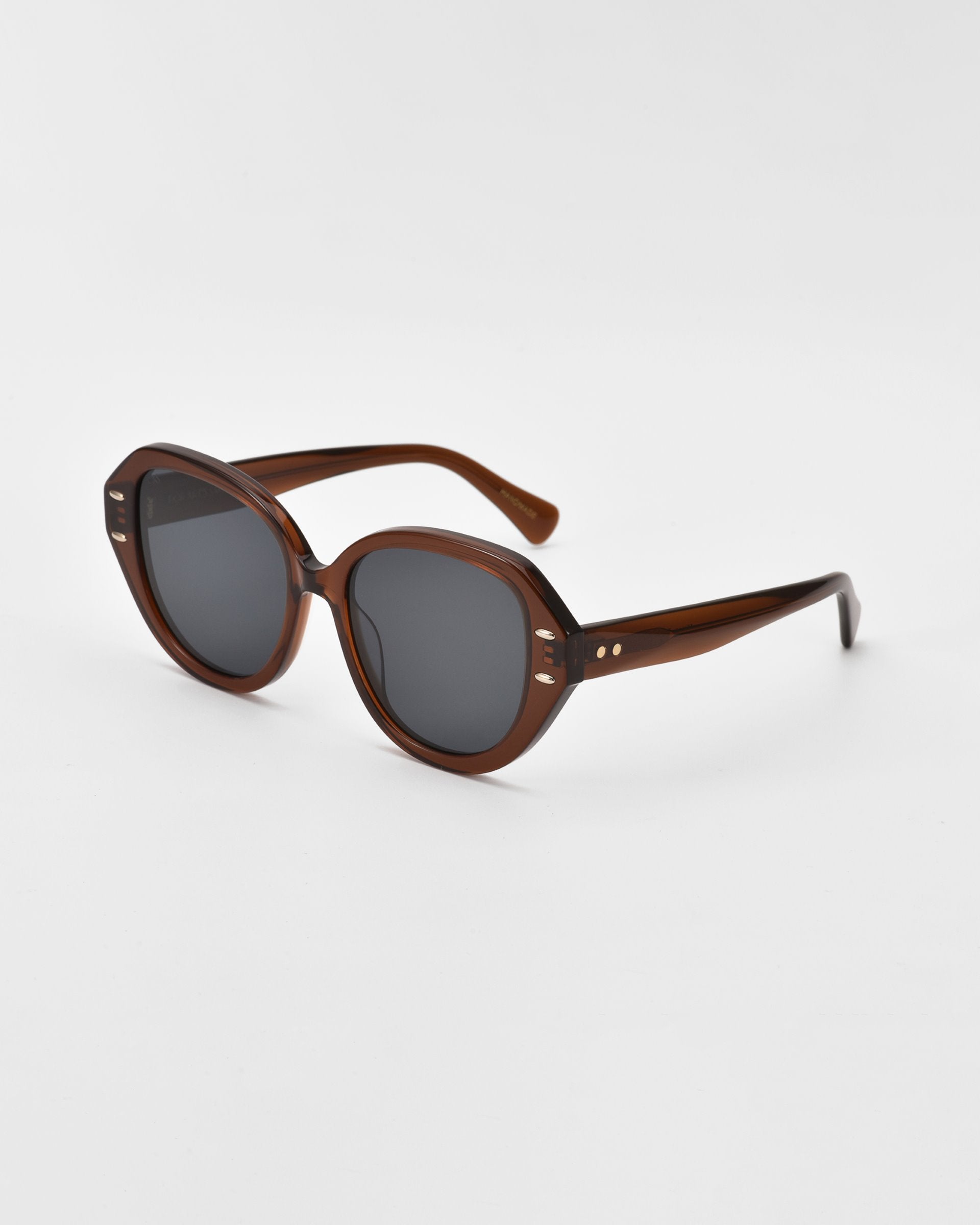 A pair of stylish dark brown Mirage cat-eye sunglasses with round frames and dark lenses, resting on a white surface. The temples feature small oval gold and silver-tone metal accents for added detail. Made from plant-based acetate, the For Art&#39;s Sake® Mirage sunglasses boast a classic yet modern design.