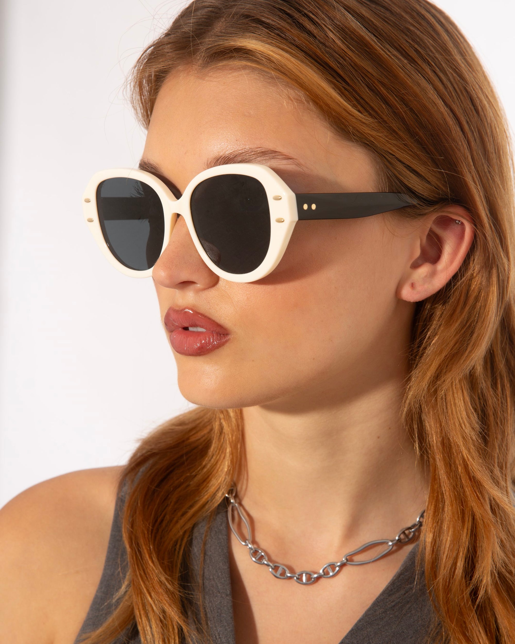 A person with long auburn hair and glossy lipstick is wearing large, round Mirage sunglasses from For Art&#39;s Sake® made of glossy acetate. They have a neutral expression and are shown against a plain background, wearing a gray top and a chunky chain necklace in gold and silver-tone metal.