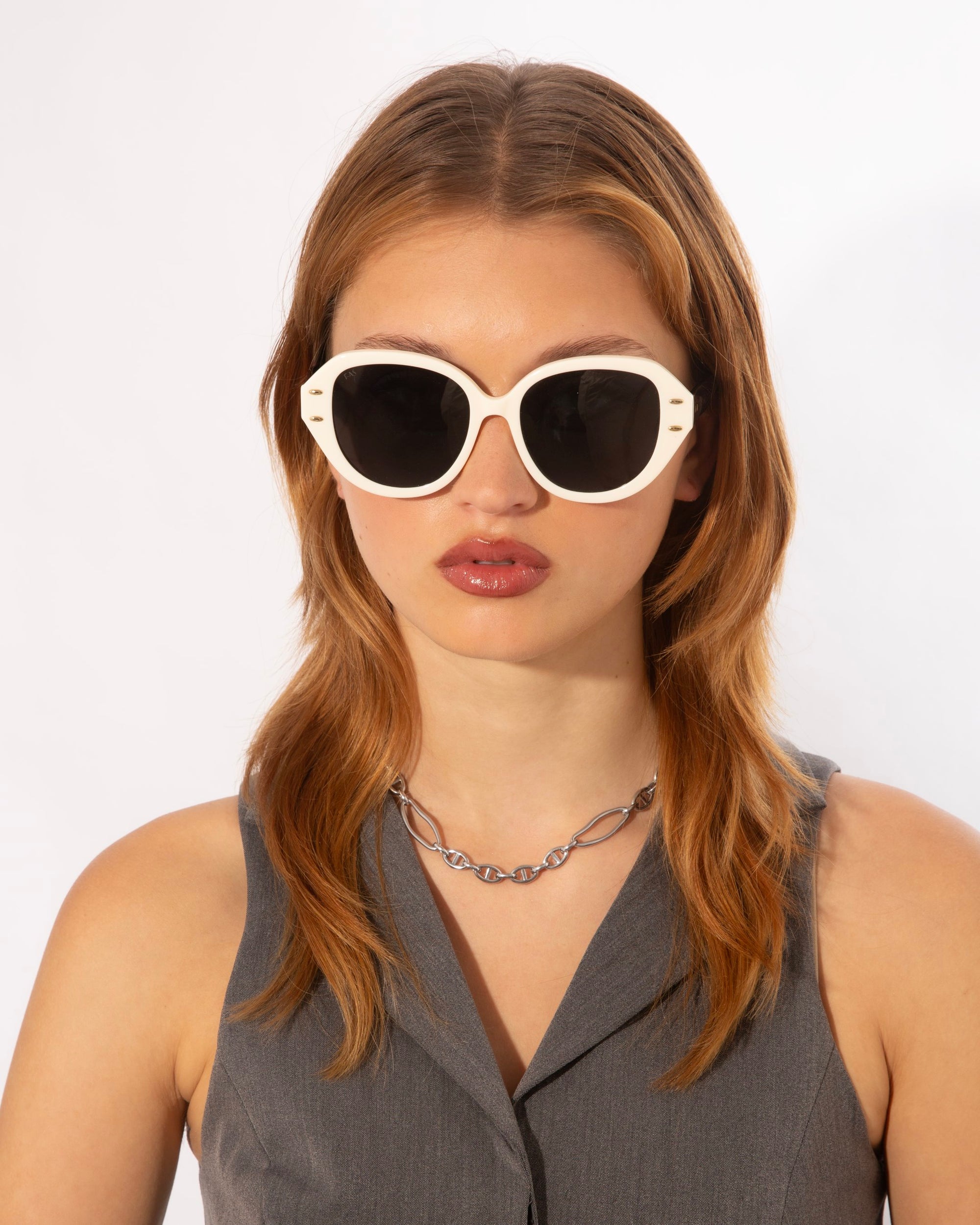A person with shoulder-length light brown hair is wearing large white-framed Mirage sunglasses from For Art&#39;s Sake® made from plant-based acetate and a sleeveless charcoal gray top. They also have a silver chain necklace. The background is plain white.