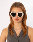 A person with shoulder-length light brown hair is wearing large white-framed Mirage sunglasses from For Art's Sake® made from plant-based acetate and a sleeveless charcoal gray top. They also have a silver chain necklace. The background is plain white.