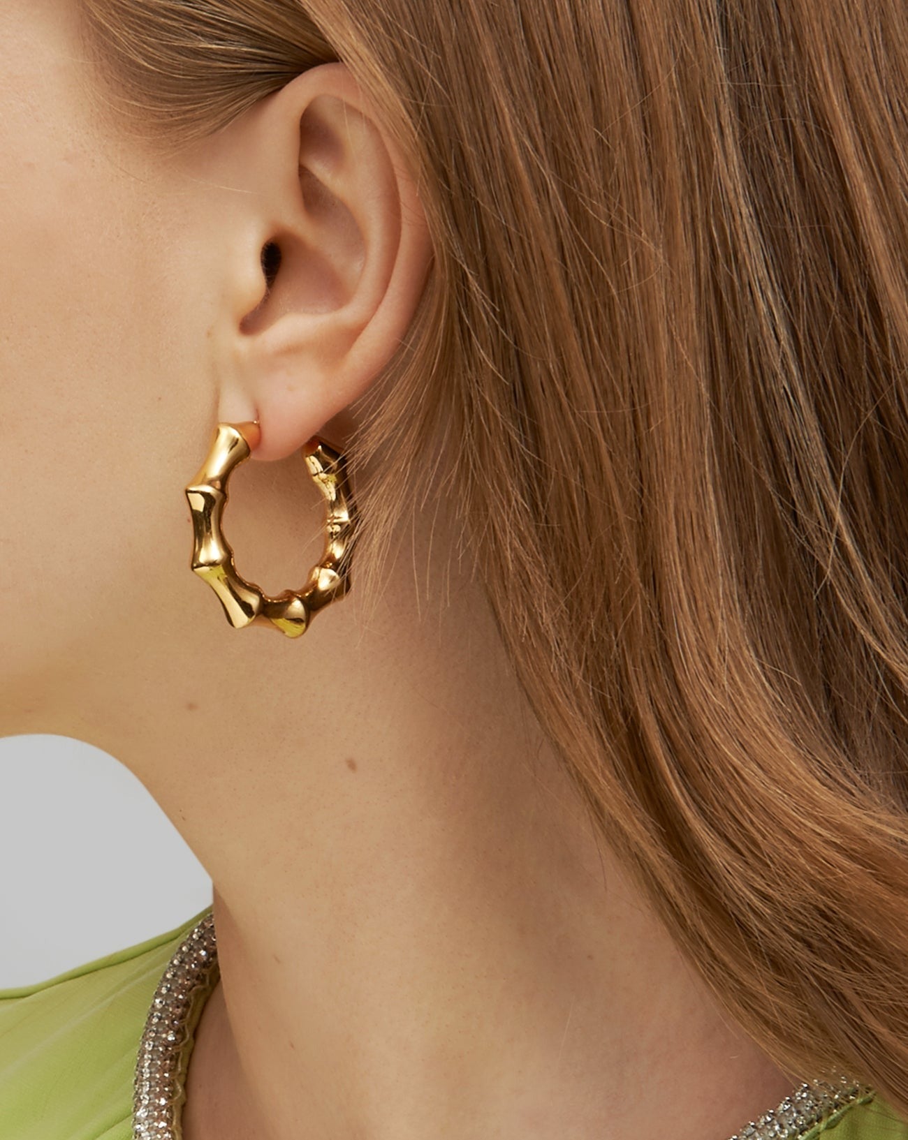 Close-up of a person with light brown hair wearing For Art&#39;s Sake® Bamboo Earrings Gold with a post-back fastening. The person is dressed in a light green top, its neckline adorned with sparkling stones. Only the ear and part of the hair and top are visible.
