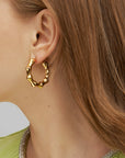 Close-up of a person with light brown hair wearing For Art's Sake® Bamboo Earrings Gold with a post-back fastening. The person is dressed in a light green top, its neckline adorned with sparkling stones. Only the ear and part of the hair and top are visible.