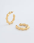 A pair of Bamboo Earrings Gold from For Art's Sake® with a unique, wavy, and textured design. Featuring a shiny finish and post-back fastening, they are displayed on a plain white background, with one standing upright and the other lying flat.
