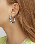 Close-up of a person wearing Bamboo Earrings Silver by For Art's Sake® with a unique, irregular design. The person has light brown hair and is dressed in a light green garment with a beaded neckline. The background is plain and light-colored.