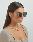 A woman with long, straight hair wearing the For Art's Sake® Birthday Cake sunglasses, which feature gold-plated frames and shatter-resistant lenses, is posed against a plain background. She is adorned with a gold necklace featuring a pendant and wearing a beige top with a square neckline. One pearl earring is visible.