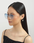 A person with long, sleek black hair is wearing the round, reflective Birthday Cake sunglasses from For Art's Sake®, which feature UV-protected, shatter-resistant lenses. They are dressed in a thin-strapped black top and have a calm expression while accessorizing with a pair of dangling pearl earrings. The background is a plain, light color.