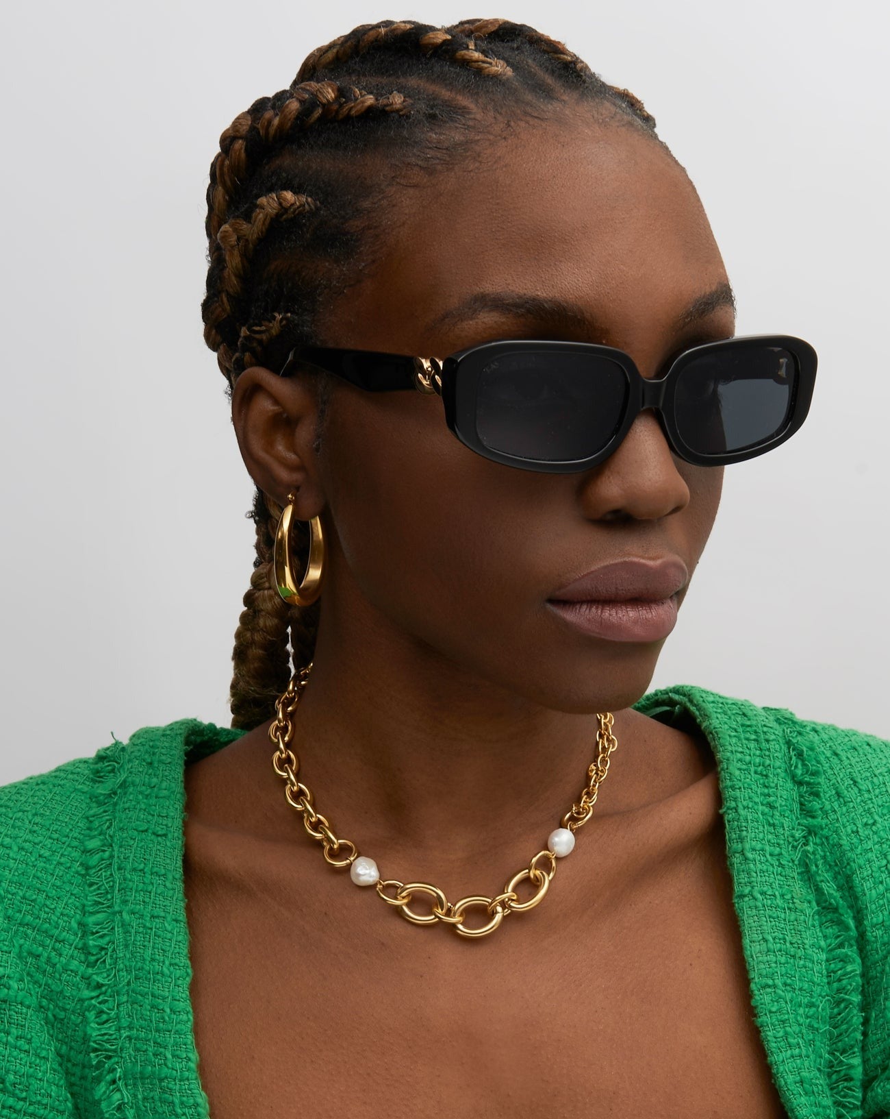 A model with dark skin and braided hair wearing the For Art&#39;s Sake Bolt black sunglasses and a green cardigan with a gold and pearl necklace. 