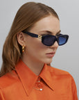 An image of a woman with Auburn hair wearing the Bolt sunglasses in Navy with an orange shirt and gold hoop earrings. 