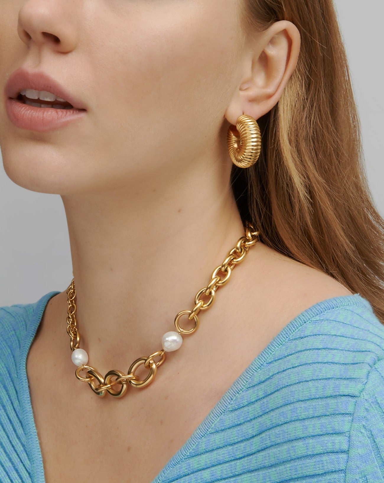 A close-up shot of a person wearing a light blue, ribbed V-neck top, Dahlia Earrings Gold by For Art's Sake®, and a chunky gold chain necklace with pearls. The background is a plain light grey, focusing attention on the jewelry and the intricate ribbed texture of the neckline.