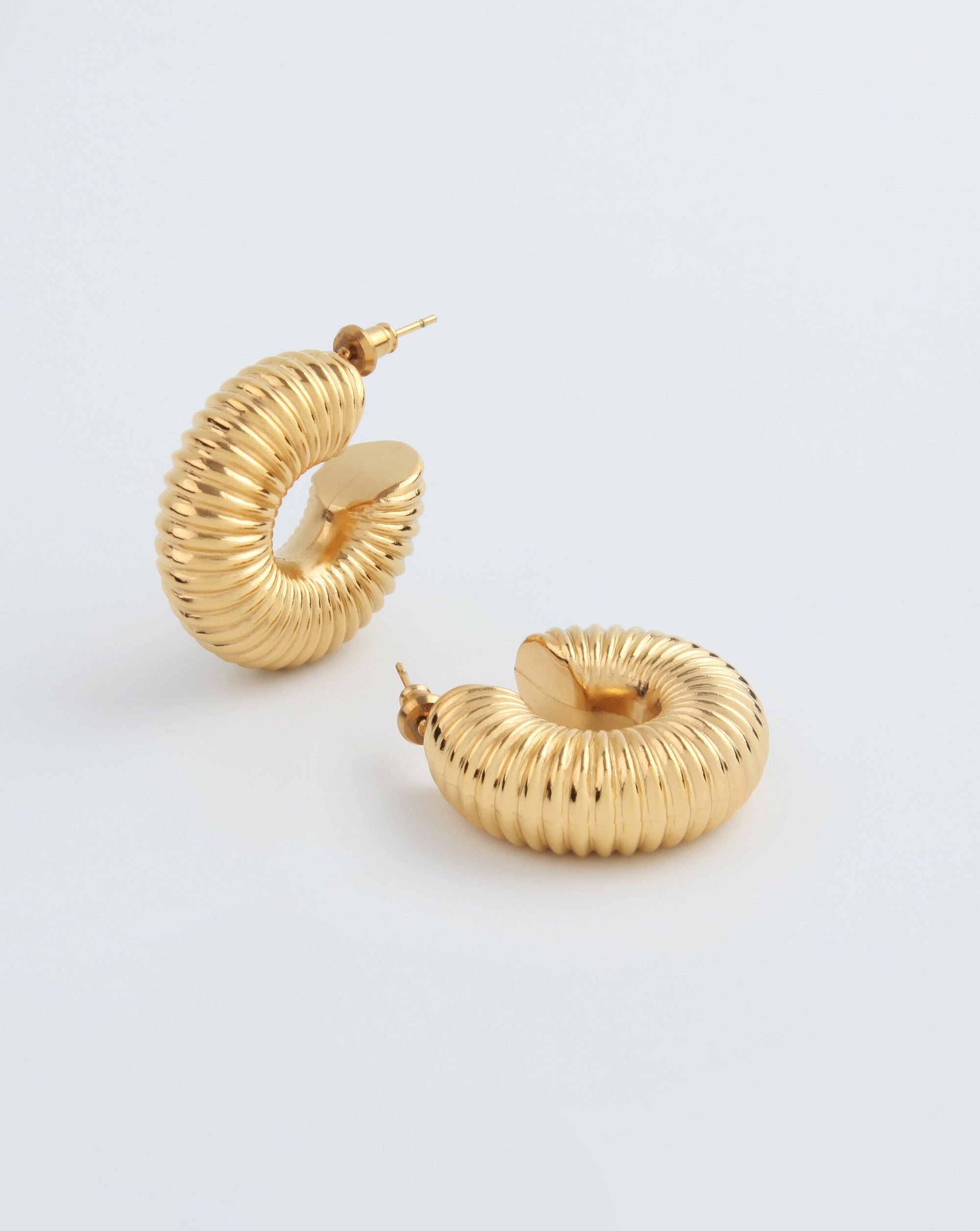 A pair of chunky tubular hoop earrings with a ribbed texture is displayed on a plain white background. One Dahlia Earrings Gold by For Art's Sake® stands upright, showcasing its circular shape, while the other lies on its side, emphasizing the intricate detailing.