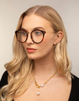 A woman with long, wavy blonde hair wears the Jackie glasses from For Art's Sake®, which are large and round with a tortoiseshell design made from plant-based acetate. She has a small ear piercing and is dressed in a black top. She accessorizes with a gold chain necklace featuring a pearl and a geometric charm. The background is plain and neutral.