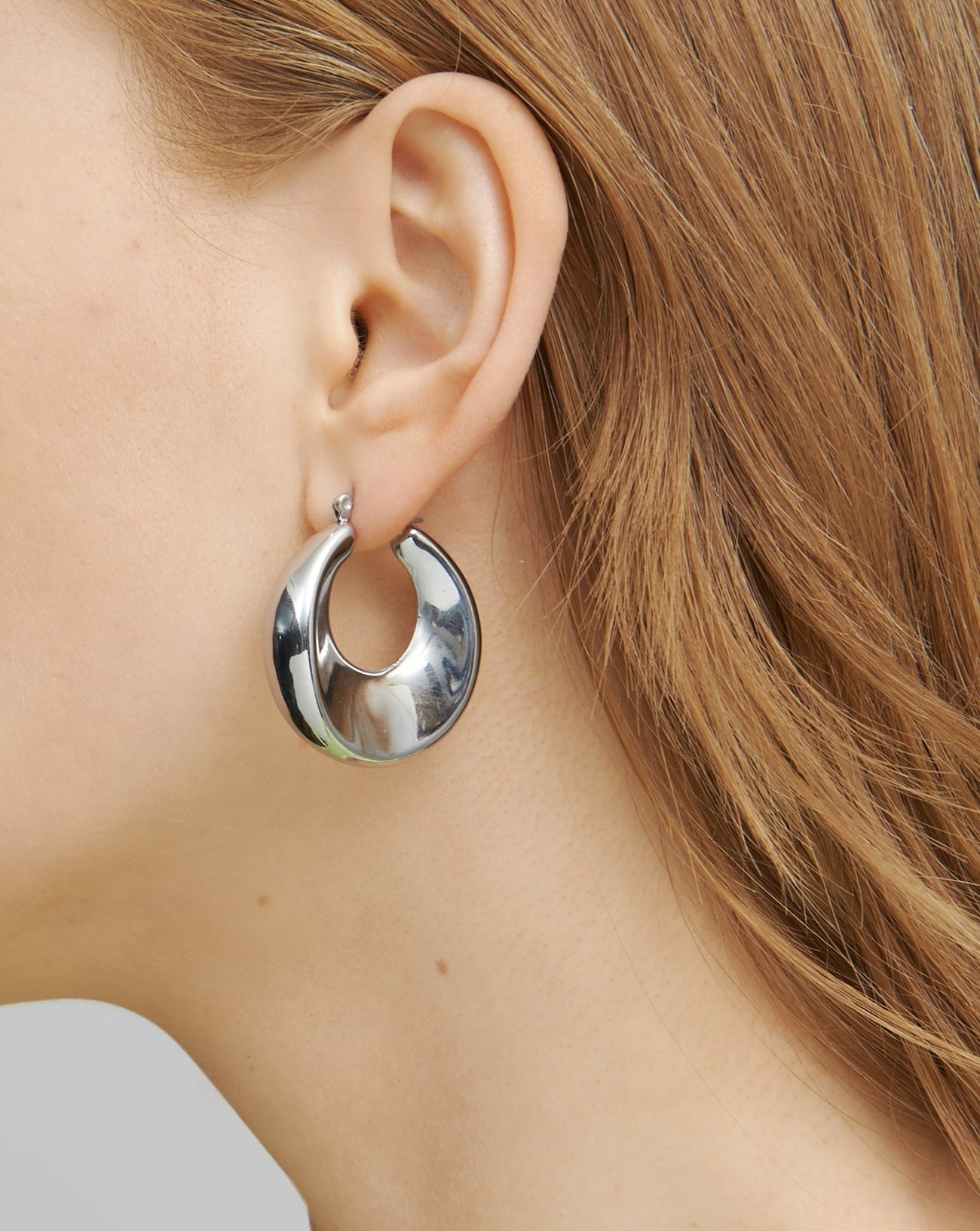 Close-up of a person with light brown hair wearing large, shiny, silver hoop earrings. The photo focuses on the ear and part of the person's hair and cheek, showcasing the distinctive design of the lightweight Moon Earrings Silver by For Art's Sake® against a plain background.
