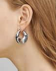 Close-up of a person with light brown hair wearing large, shiny, silver hoop earrings. The photo focuses on the ear and part of the person's hair and cheek, showcasing the distinctive design of the lightweight Moon Earrings Silver by For Art's Sake® against a plain background.