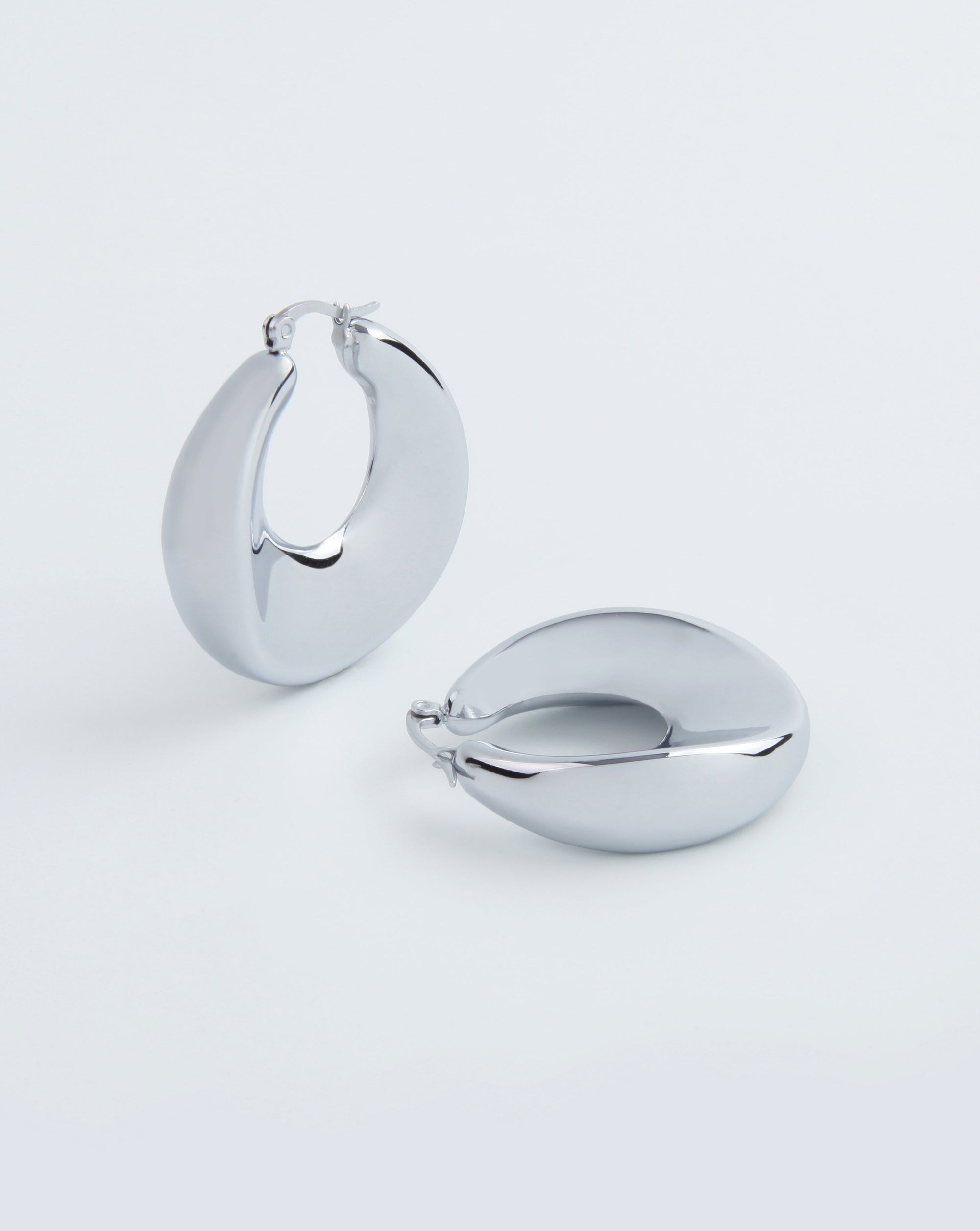 A pair of shiny, smooth, 18kt gold-plated hoop earrings are displayed against a plain white background. One earring stands upright, while the other lies flat, showcasing their sleek and polished design. Despite their luxurious appearance, these Moon Earrings Silver by For Art's Sake® are remarkably lightweight.