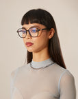 A woman with straight black hair and black cat-eye framed optical glasses, wearing a sheer, ribbed top and a chain necklace.