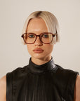 A woman with a short, blunt bob haircut wearing the Azure brown optical glasses by For Art’s Sake and a black sleeveless turtleneck, giving a serious look toward the camera against a neutral background.