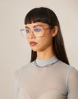 A woman with straight black hair and clear-frame optical glasses, wearing a sheer, ribbed top and a chain necklace.