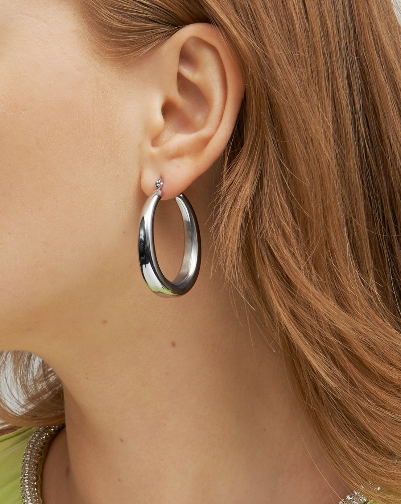 Close-up of a person wearing a large, smooth, For Art's Sake® Oval Earrings Silver with a small diamond accent. The person's ear and part of their reddish-brown hair are visible, and they are wearing a light green top with a beaded collar.