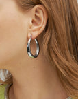 Close-up of a person wearing a large, smooth, For Art's Sake® Oval Earrings Silver with a small diamond accent. The person's ear and part of their reddish-brown hair are visible, and they are wearing a light green top with a beaded collar.