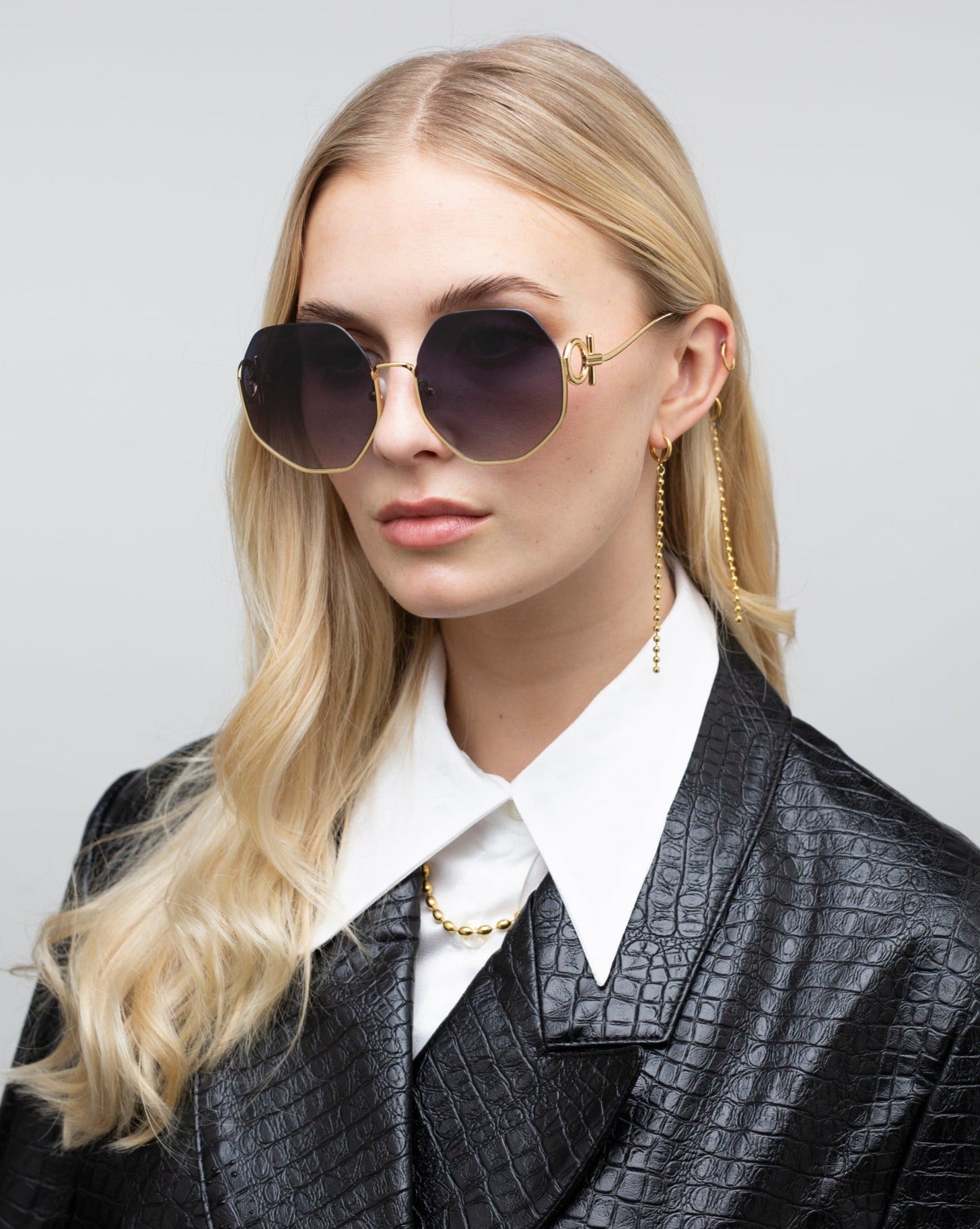 A woman with long blonde hair wearing large, round For Art's Sake® Palace sunglasses that are UVA & UVB-protected and a black, textured leather jacket. She has a white collared shirt underneath and is accessorized with a dangling earring and an 18-karat gold-plated chain necklace. The background is plain and light-colored.