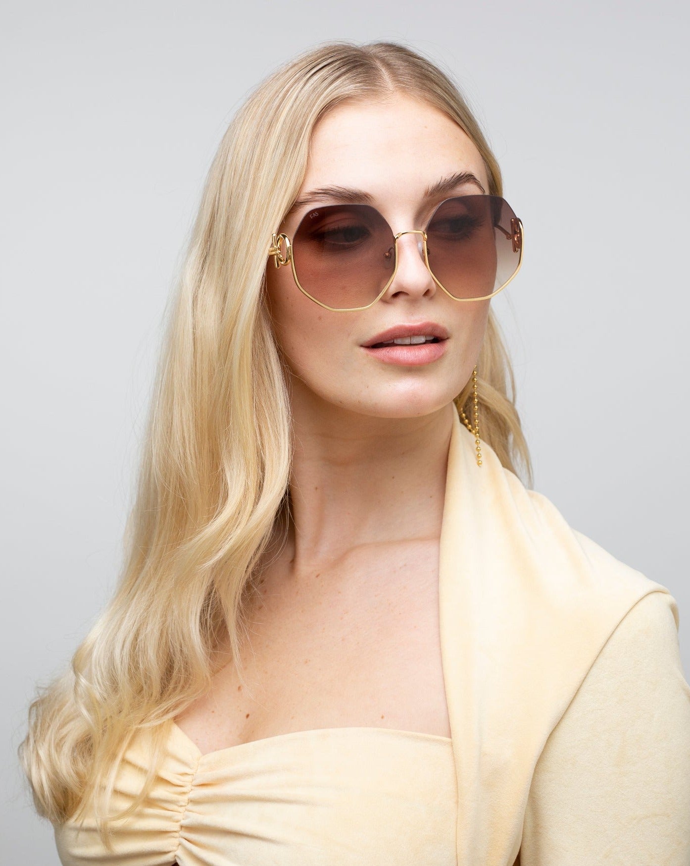 A woman with long, blonde hair is wearing large, stylish For Art&#39;s Sake® Palace sunglasses with 18-karat gold-plated frames and brown-tinted, UVA &amp; UVB-protected lenses. She is dressed in a light beige outfit and is looking slightly to the side with a neutral expression. The background is plain and light gray.