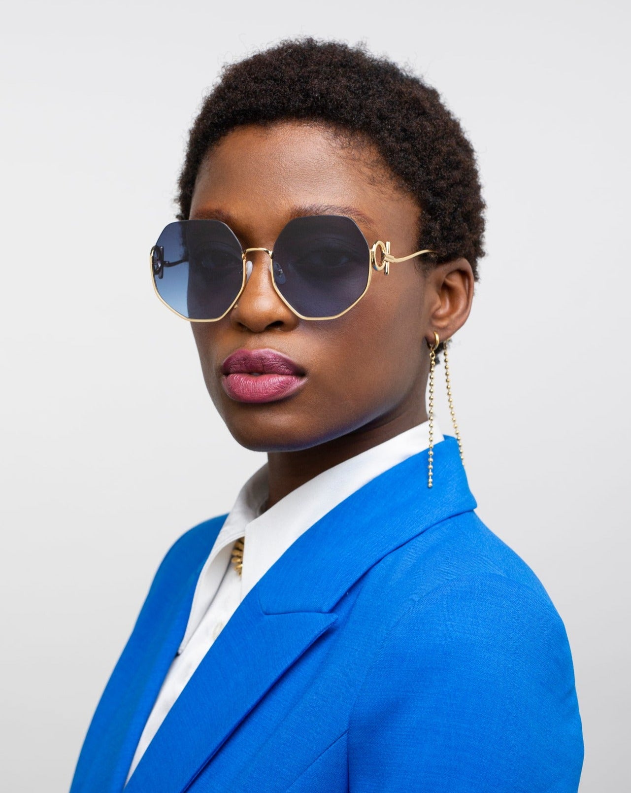 A person wearing octagonal navy blue sunglasses with UVA & UVB protection from For Art's Sake® called Palace, a blue blazer over a white shirt, and an earring with multiple 18-karat gold-plated chains running down from it. The person has short curly black hair and is standing against a plain light grey background.
