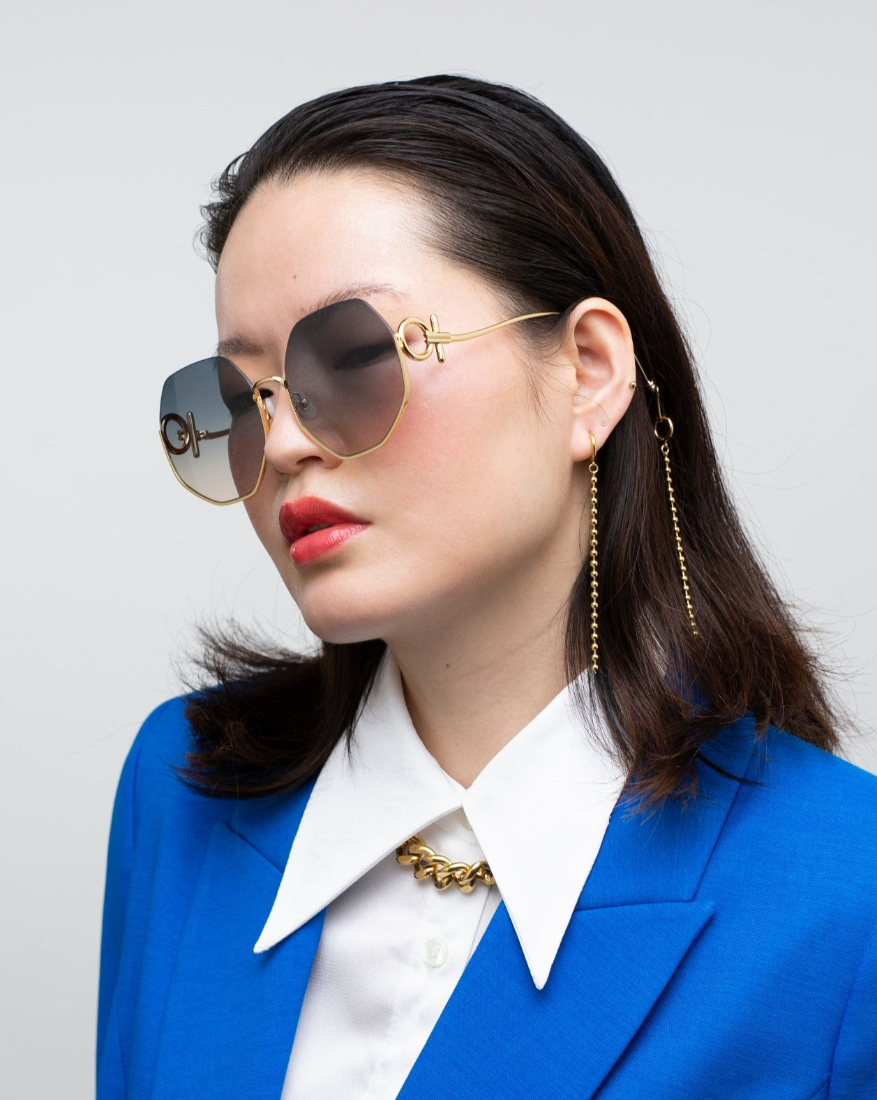 A person with straight, dark hair is wearing large, UVA &amp; UVB-protected For Art&#39;s Sake® Palace gold-rimmed sunglasses with gradient lenses. They are dressed in a blue blazer over a white collared shirt and accessorized with 18-karat gold-plated chain earrings and necklace. The background is a solid light gray.
