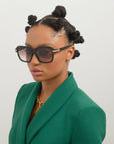 A person wearing Shadyside sunglasses by For Art's Sake®, handmade from acetate and offering UVA & UVB protection, alongside a green blazer paired with an 18-karat gold-plated necklace. Their hair is styled in multiple small buns against a plain white background.