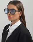 Sporting Shadyside handmade acetate sunglasses with large, rectangular blue-tinted lenses from For Art's Sake® and 18-karat gold-plated hoop earrings, a woman with long, straight hair is dressed in a black blazer over a white collared shirt. She gazes off to the side against a plain, light gray background.